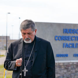Bishop in black holds a rosary outside Hudson Correctional Facility sign in Hudson, Colorado, looking somber.