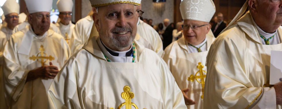 Catholic bishops in white robes and miters walk indoors in procession, smiling in a warmly lit hall.