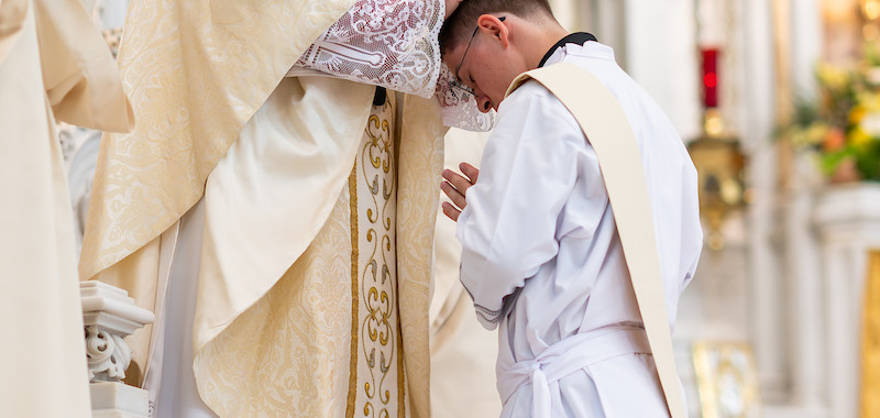 A bishop in ornate robes prays over a kneeling young man in a white robe inside a cathedral. The setting is ornate and solemn.