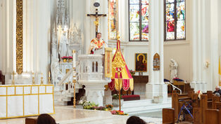 A priest in ornate vestments speaks from a pulpit in a bright church, with stained glass and religious statues in the background.