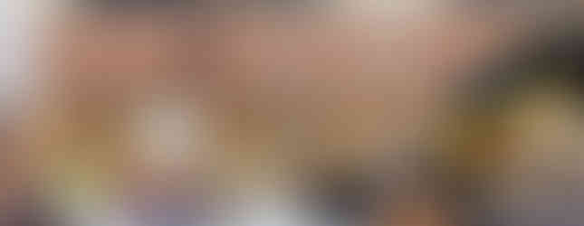 Audience watches a nun speaking into a microphone at an outdoor event. Children in yellow helmets and safety vests sit nearby. Construction equipment visible.