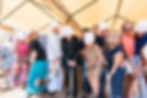 Group of people in hard hats and dress clothes shovel dirt at a ceremonial groundbreaking under a white tent.