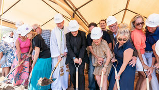 Group of people in hard hats and dress clothes shovel dirt at a ceremonial groundbreaking under a white tent.
