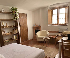 A practical ground-floor studio apartment with terracotta floors and white walls. The space includes a designated study nook at the entrance, a cozy sleeping area, and a functional kitchenette. The unit opens onto a quiet, traditional paved courtyard.