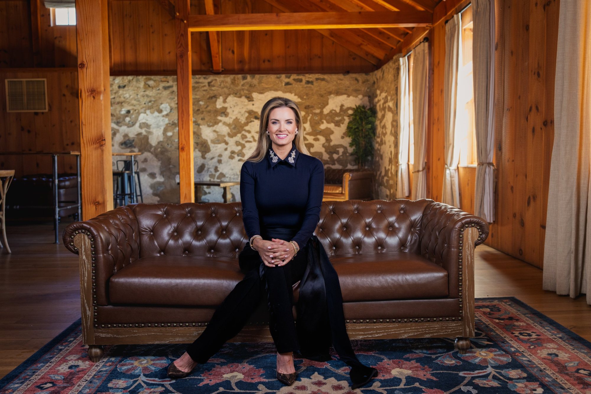Jillian smiling on a leather couch in a sunlit barn, in a black top with a jeweled collar