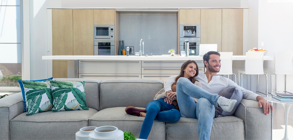 Smiling couple sits on a gray sofa with tropical cushions in a bright kitchen. Light wood cabinets and modern decor create a cozy atmosphere.