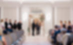 Bride and groom stand under floral arch in elegant room, surrounded by bridal party and guests, during a wedding ceremony.
