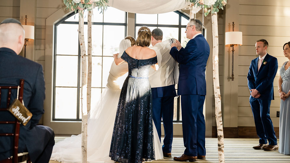 Wedding ceremony under floral chuppah. Couple embraces as others stand nearby. Elegant attire, bright indoor setting, emotional ambiance.