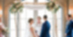 Bride and groom stand under floral arch in indoor ceremony. Bridesmaid holds bouquet. Officiant reads. Soft natural light filters in.