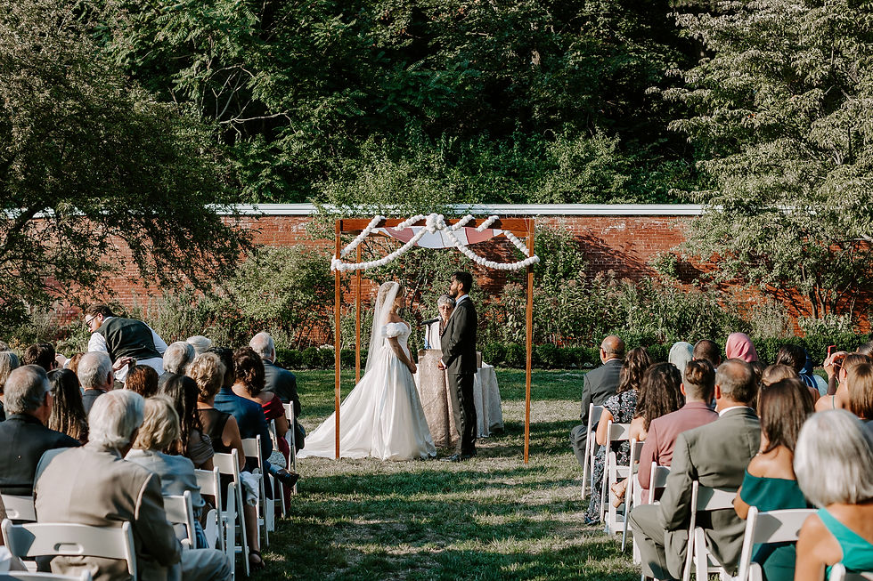 Bride and groom stand under a wood chuppah during their wedding ceremony, exchanging vows before a seated audience.