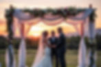 Bride and groom under floral chuppah at sunset, exchanging vows with officiant. Romantic outdoor setting, soft pink and white flowers.