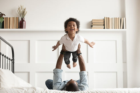 Happy funny african american kid boy flying in fathers arms looking at camera in bedroom,