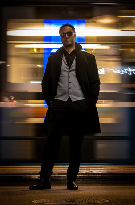 A portrait of an African-American man wearing gold sunglasses, a black turtleneck sweater and coat, standing at a subway station at night.