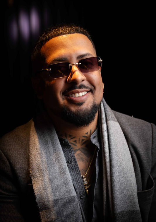 Portrait of a smiling man wearing sunglasses and a grey scarf against a dark background.