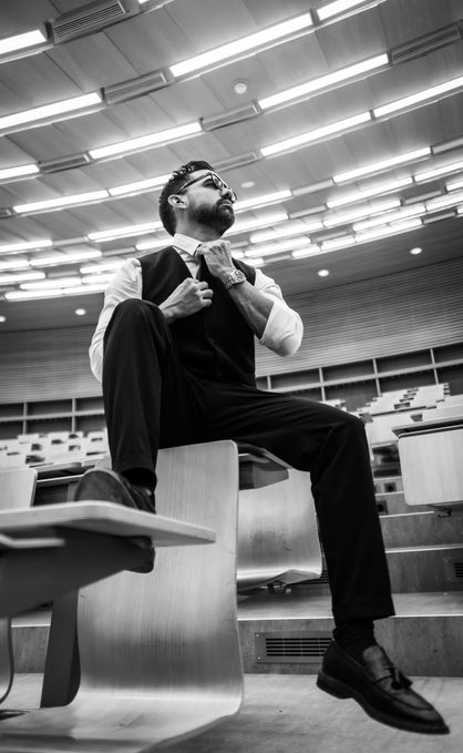 A black and white portrait of a man in a suit looking upwards in a lecture hall with wooden rows of chairs.