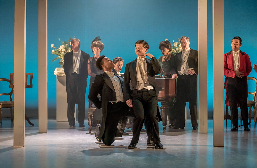 The cast of The Land of Might-Have-Been gather around a piano within a stately home setting