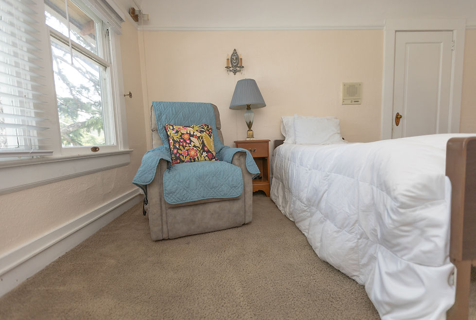 "Private resident bedroom with a single bed, upholstered chair, bedside table, and natural light from a large window."
