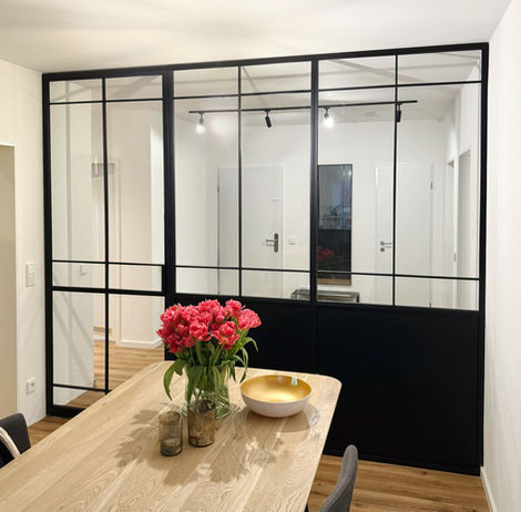 Glastrennwand mit Pivot-Tür im schwarzen Stahlrahmen RAL 9005 und unterem Metallteil, die den Flur vom Esszimmer in einem modernen Interieur trennt.