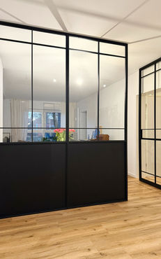 Glastrennwand mit Pivot-Tür im schwarzen Stahlrahmen RAL 9005 und unterem Metallteil, die den Flur vom Esszimmer in einem modernen Interieur trennt.