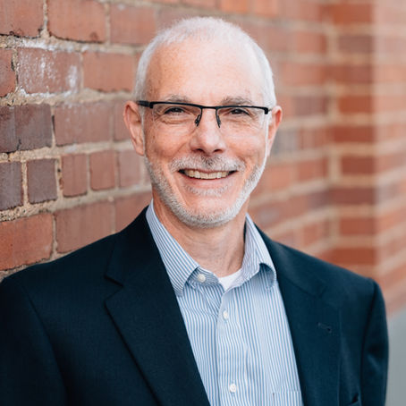 Smiling man with glasses in a suit, leaning against a brick wall. The background is a warm-toned brick pattern, creating a friendly mood.