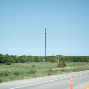 Cell tower in a rural field, surrounded by hay bales and trees under a clear blue sky. Road with orange cones in the foreground.