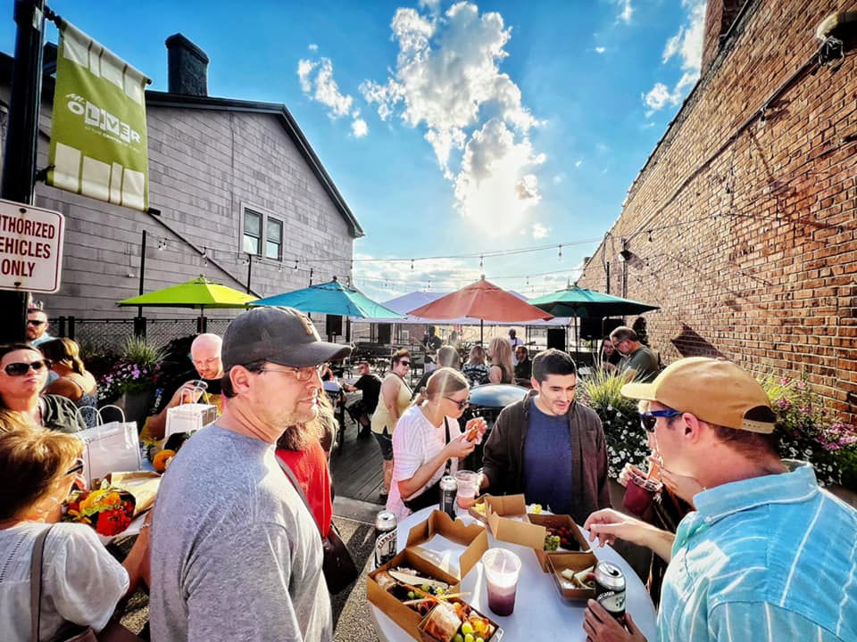 People enjoying food and drinks at an outdoor event with colorful umbrellas, brick walls, and a bright sky. Energetic and lively mood.