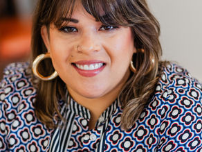 Woman smiling in a patterned blouse with earrings. Blurred orange and gray background, conveying a friendly, warm mood.