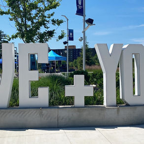 Large "WE+YOU" letters in a park setting with green plants, blue sky, colorful tents, and people in background; vibrant and inviting.