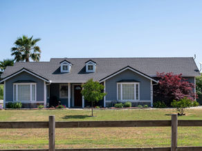 Blue house with gabled roof and white trim, surrounded by green lawn and trees. Clear blue sky and wooden fence in the foreground.