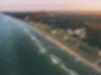 Aerial view of a scenic coastline with lush forests meeting the ocean. Gentle waves, sandy dunes, and a sunset sky create a tranquil scene.