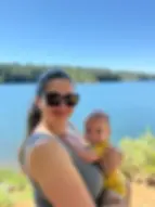 Priya holding her baby son in front of the beautiful Paradise Lake, with
green trees on the far shore and a clear blue sky.