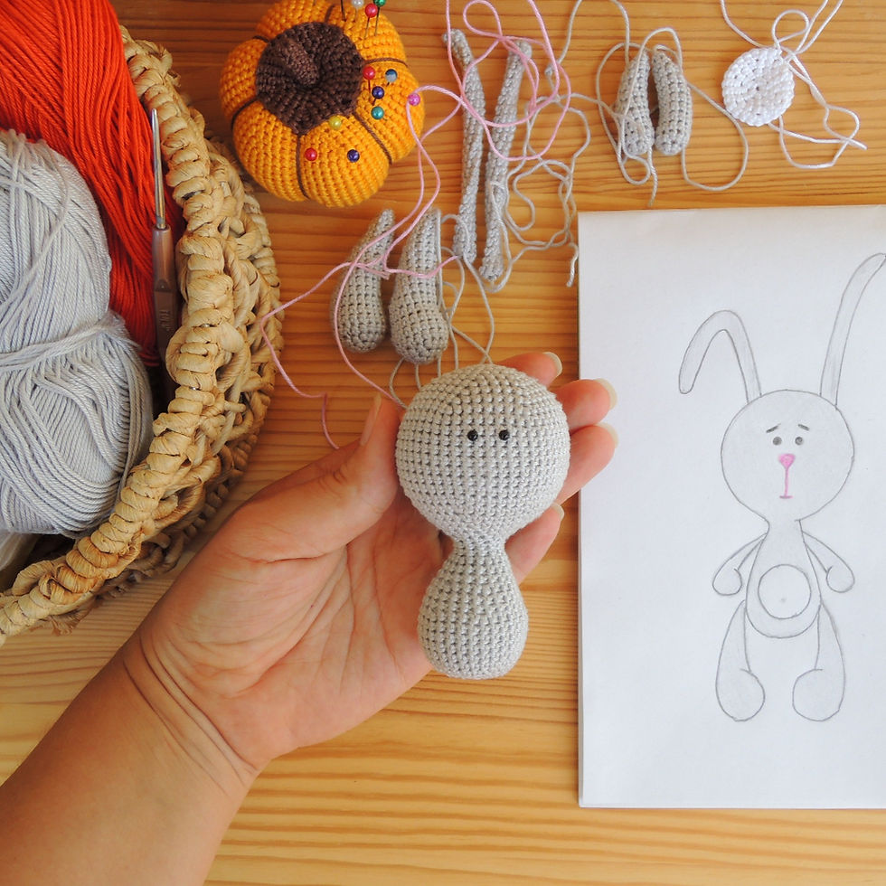 Hand holding a gray crochet bunny head, surrounded by yarn, crochet tools, and a sketch of a bunny design on a wooden table.