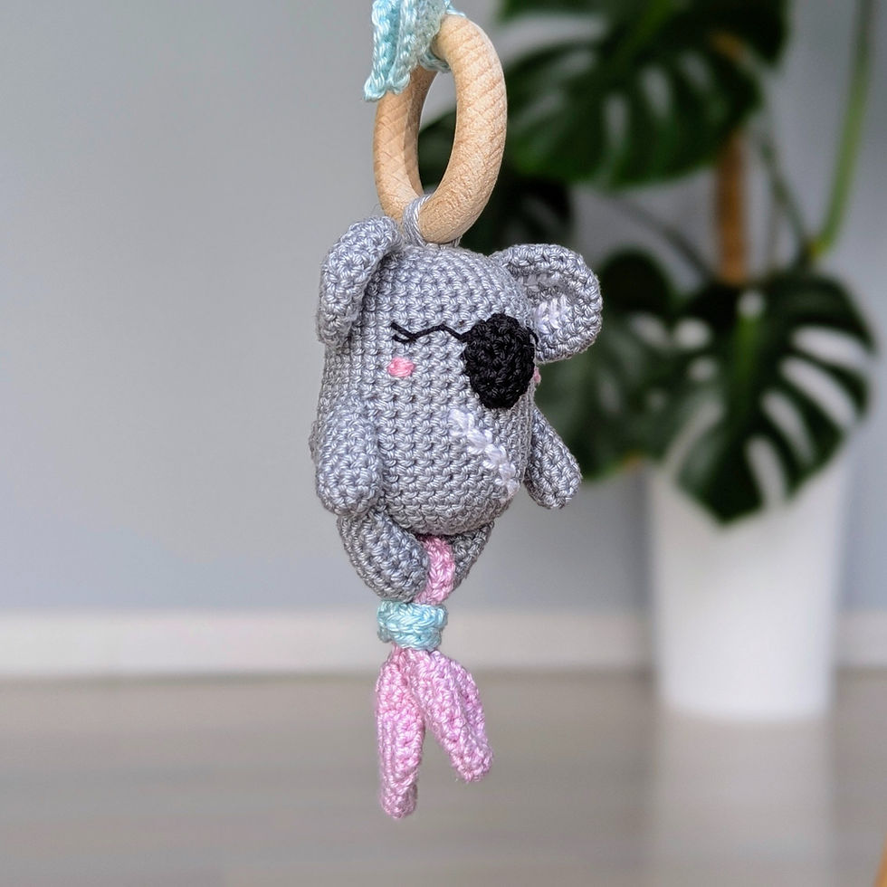 Thumbnail: Crocheted koala rattle with sewn-on nose and embroidered smiling eyes and rosy cheeks, and with small lilac leaves below