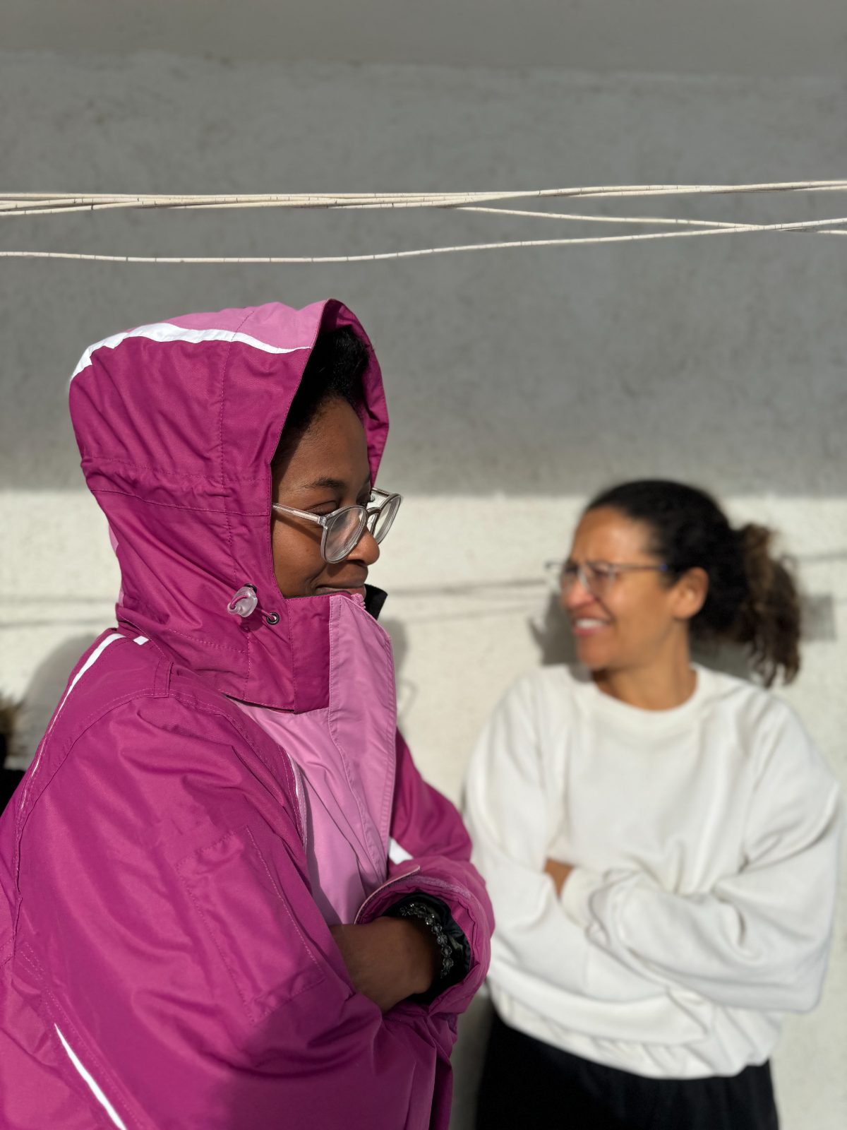 A woman in a pink hooded jacket smiling in the sun, another woman behind her