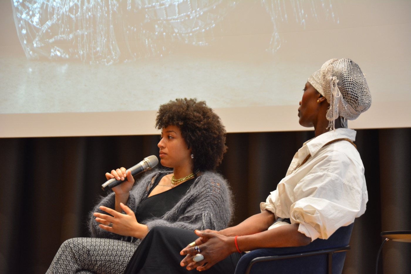 Christin Khaukha speaking into a microphone on stage, next to a guest in a white shirt