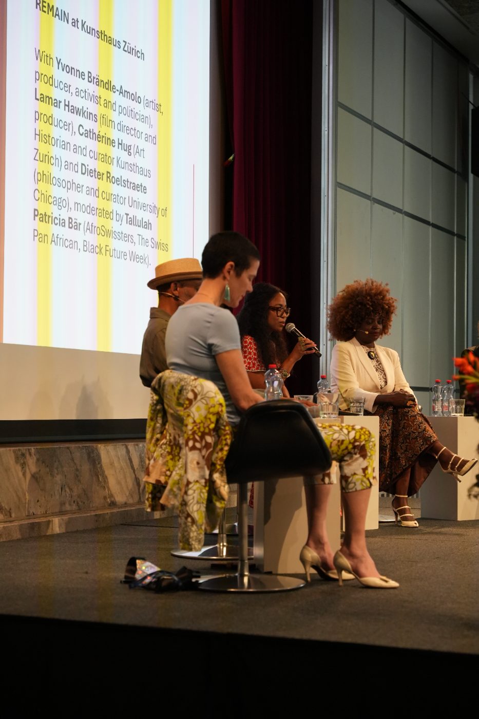 Tallulah Patricia Bär speaking on stage, next to Lamar Hawkins and Yvonne Apiyo Brändle-Amolo