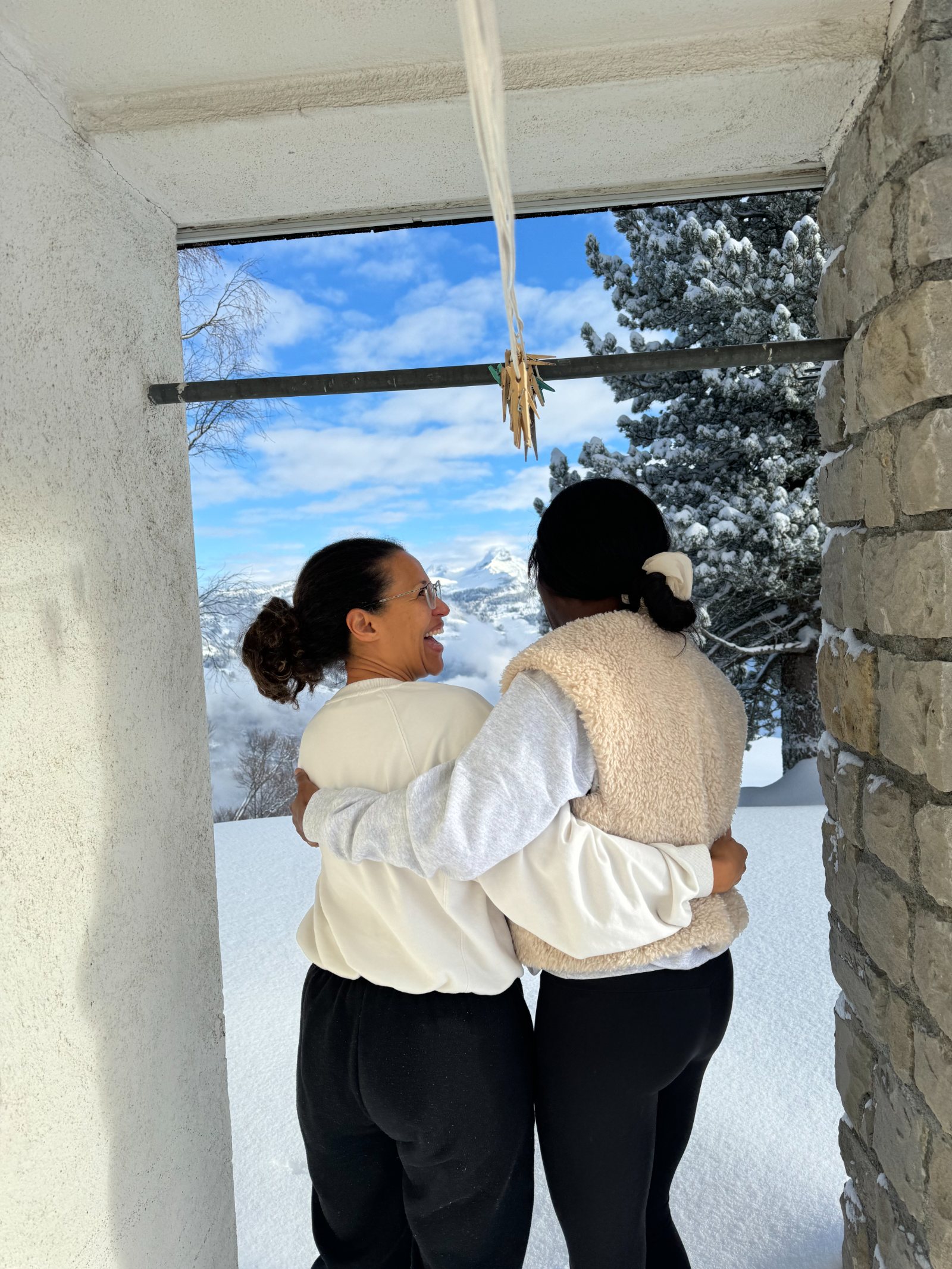 Two women hugging in a doorway, snow and fir trees outside