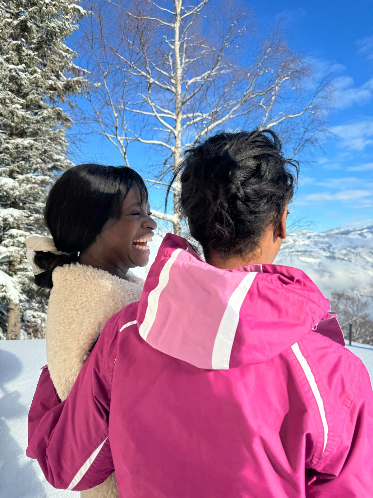 A woman laughing beside another woman in a pink jacket, with snowy trees and blue sky behind them