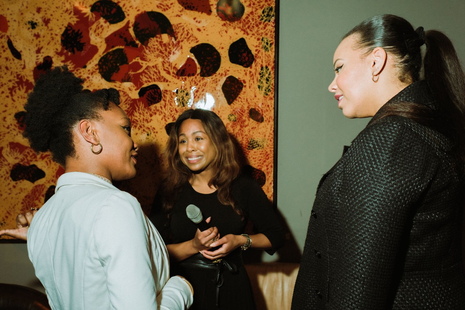 Three women in conversation in front of a large painting, one holding a microphone