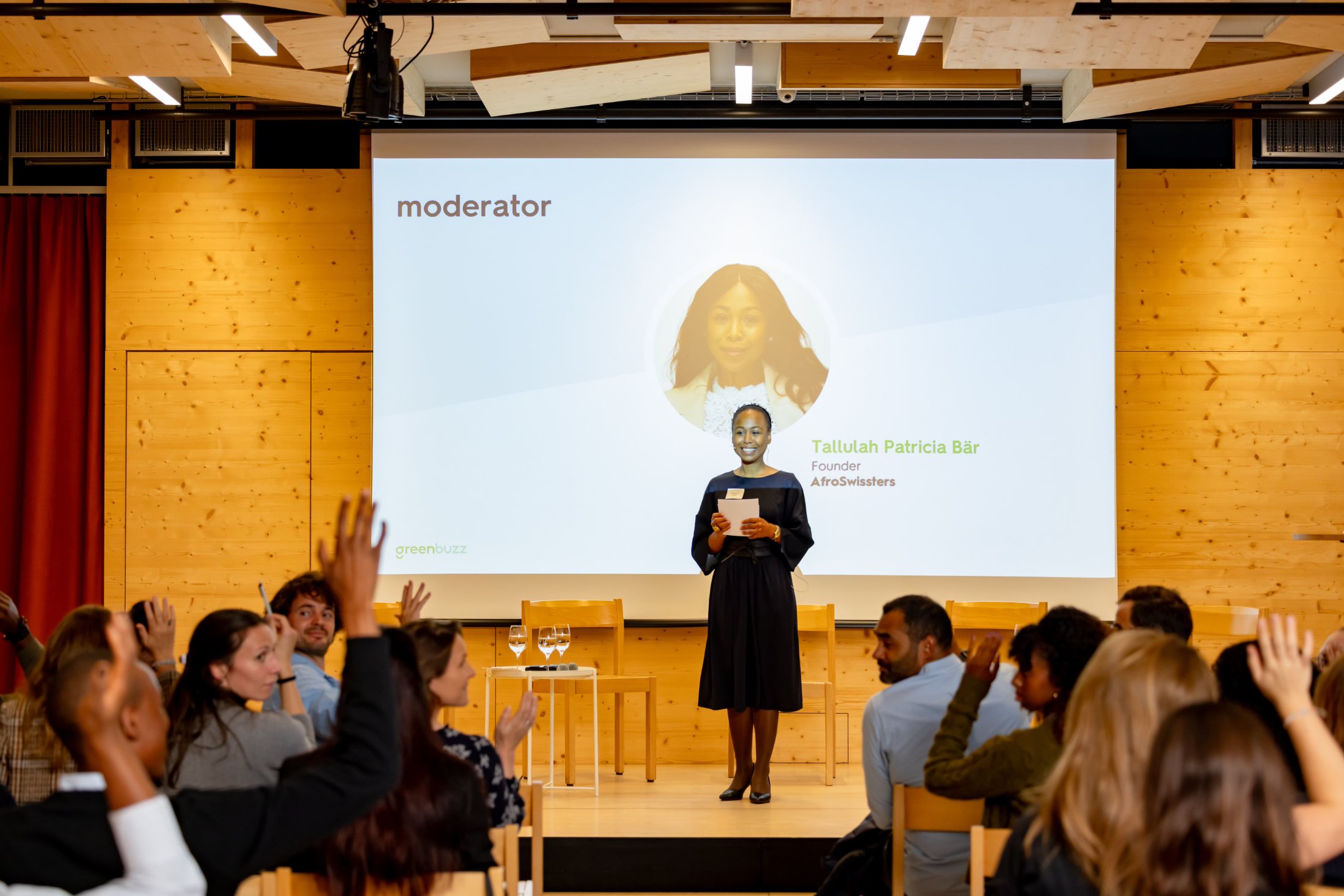 Tallulah Patricia Bär on stage as moderator at a Greenbuzz event, the audience raising hands