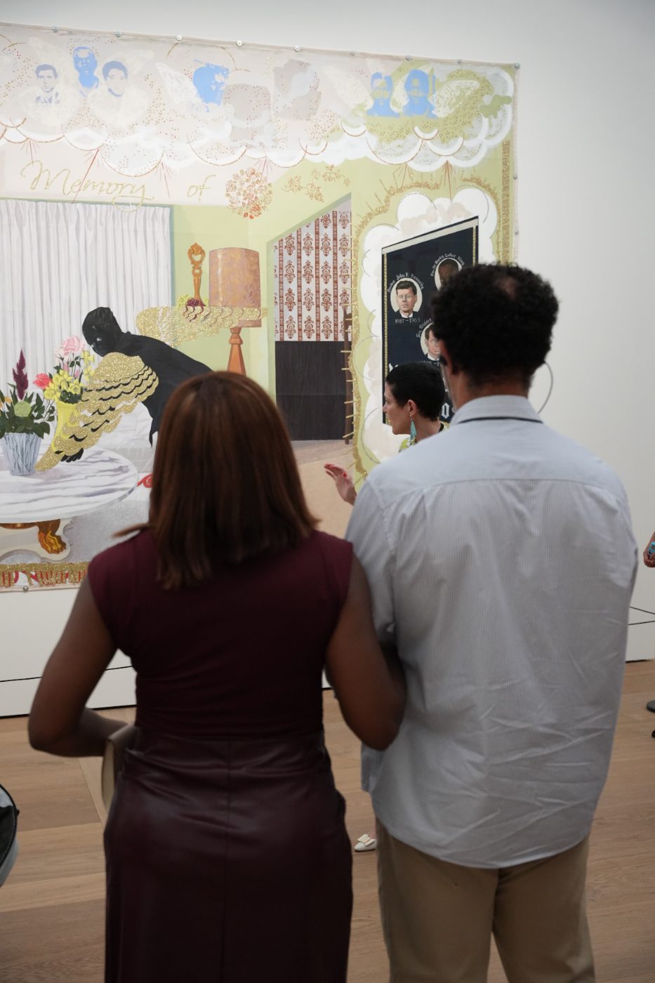Two guests in front of a painting with the words “Memory of”, the curator in the background