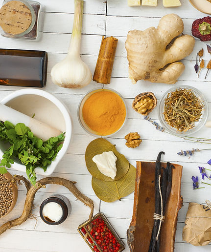 Assortment of herbal medicine ingredients on a white wooden surface, including fresh herbs, powders, nuts, vanilla pods, red berries, and dried roots for natural health remedies