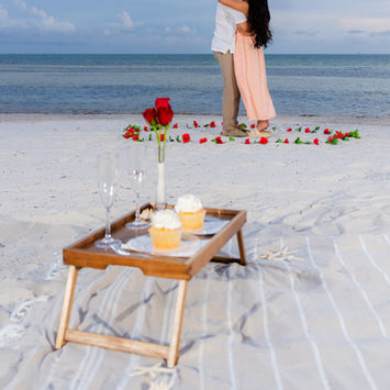 A beach proposal is taking place near this proposal picnic setup with dessert and champagne