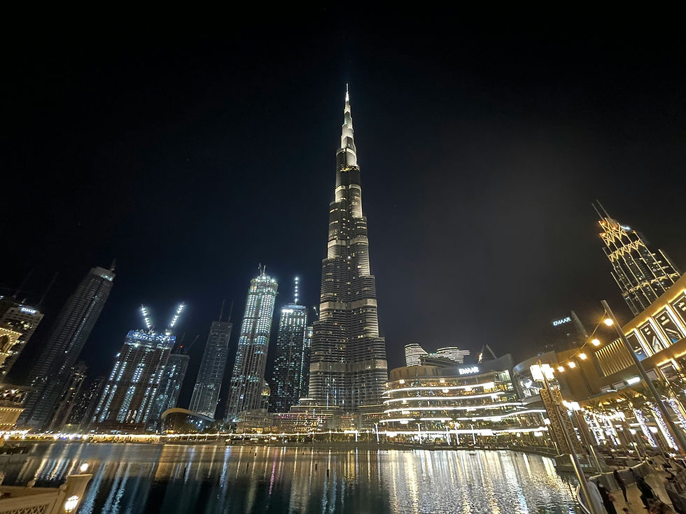 Night view of a city skyline with a tall, illuminated skyscraper reflecting in a calm water body. Lights from nearby buildings glow brightly.