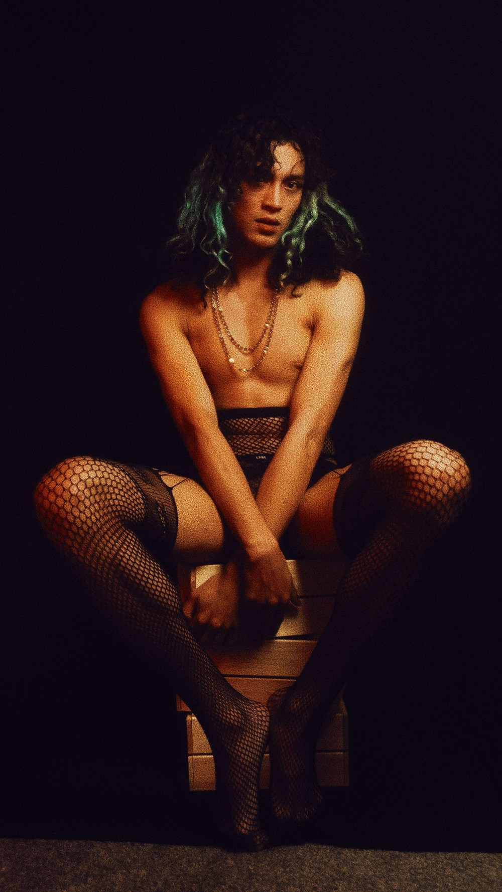 Tom Issac with blue streaks in his hair, posed wearing fishnet tights with gold chain necklaces around his neck.