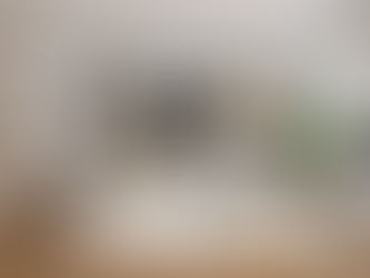 Sala de estar moderna e luminosa, com sofá branco em L, almofadas decorativas, mesa de centro com tampo em mármore e base dourada, estante embutida com livros e objetos decorativos, grande quadro abstrato em preto e branco, candeeiro de teto em fibras naturais, planta em vaso ao lado de móvel branco com televisão e cozinha aberta com bancos altos de madeira.