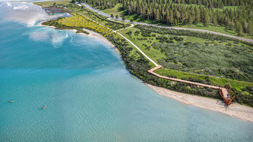 Uma paisagem costeira deslumbrante na Lagoa de Óbidos, captada de um ângulo elevado. A imagem documenta a reabilitação do Cais Palafítico da Barrosa e a criação de um novo observatório de aves. Vê-se um passadiço de madeira que se estende ao longo da encosta, o cais a avançar sobre a água, e a vibrante cor azul-turquesa da lagoa, contrastando com o verde da vegetação e da floresta.