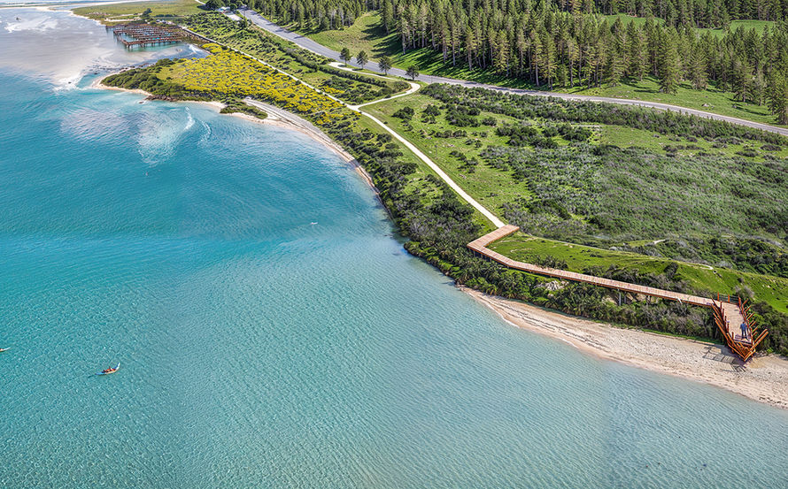 Uma paisagem costeira deslumbrante na Lagoa de Óbidos, captada de um ângulo elevado. A imagem documenta a reabilitação do Cais Palafítico da Barrosa e a criação de um novo observatório de aves. Vê-se um passadiço de madeira que se estende ao longo da encosta, o cais a avançar sobre a água, e a vibrante cor azul-turquesa da lagoa, contrastando com o verde da vegetação e da floresta.