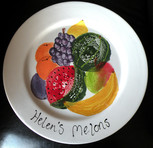 Boob prints on a plate, the prints have been turned into melons with the wording 'Helen's melons' beneath them, there is other fruit painted around the prints such as a banana and grapes.