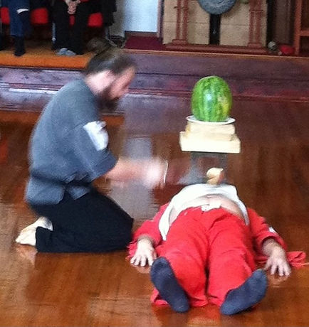 Zen Maste Sebastian Rizzon using a sword to cut an apple in half wthat is sitting on another persons stomach